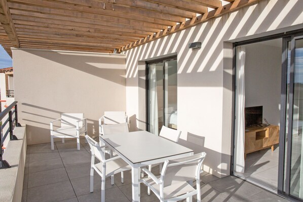 A covered outdoor patio with a white table and chairs, a wooden ceiling, and a sliding glass door leading to an interior space with a wooden cabinet and a television. - Résidence Premium Pierre & Vacances Horizon Golf (Saint-Cyprien)