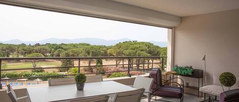 A balcony with a table and chairs, a potted plant, and a view of a green landscape with trees and mountains.
