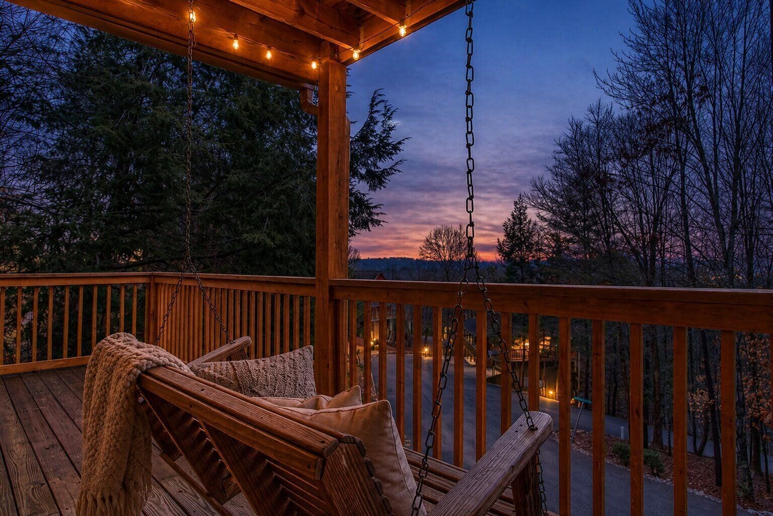A wooden deck with a swing chair and a blanket, overlooking a road and trees.