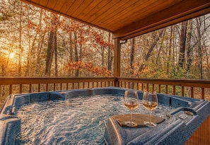A hot tub with two glasses of wine on a wooden deck with a forest view.