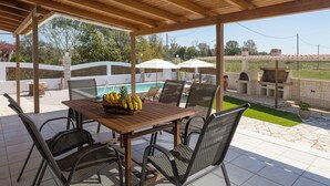An outdoor patio with a wooden table and wicker chairs, a pool, and a barbecue area.