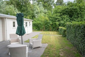 Eine Terrasse mit Korbmöbeln und einem grünen Sonnenschirm, ein weißes Haus und ein gepflegter Rasen mit einer Hecke.
