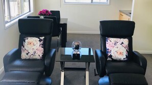 Een kamer met twee zwarte leren fauteuils met bloemkussens, een glazen bijzettafeltje met een decoratief object, en een raam met uitzicht op bomen en een hek.