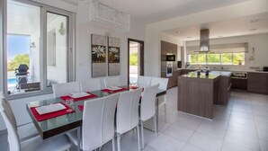 Une cuisine moderne dotée d’un îlot central, d’un coin repas avec des chaises blanches et une table en verre, et offrant une vue sur l’extérieur.