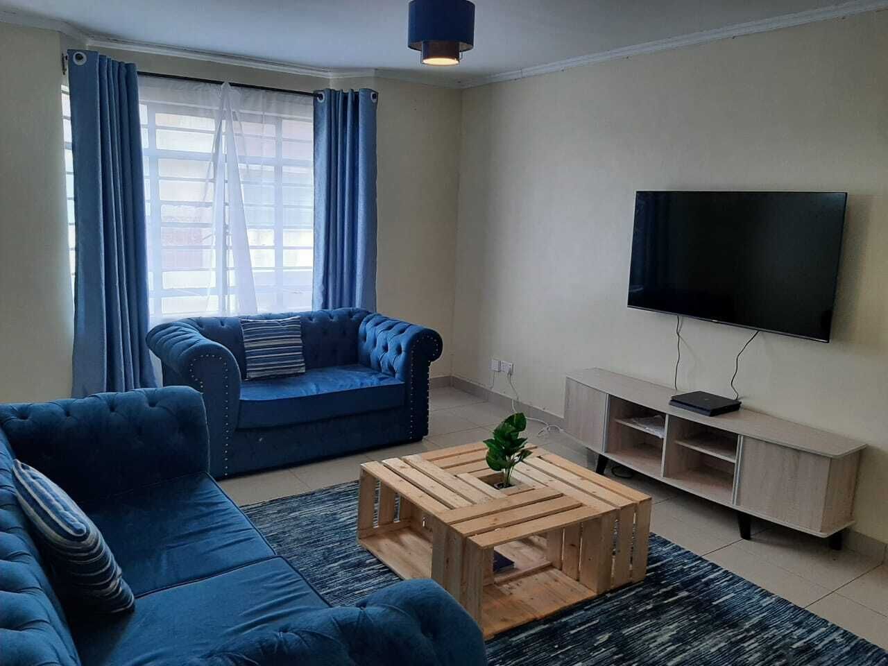 Una sala de estar con un sofá azul, una mesa de centro de madera, un televisor sobre un mueble bajo y una ventana con cortinas.