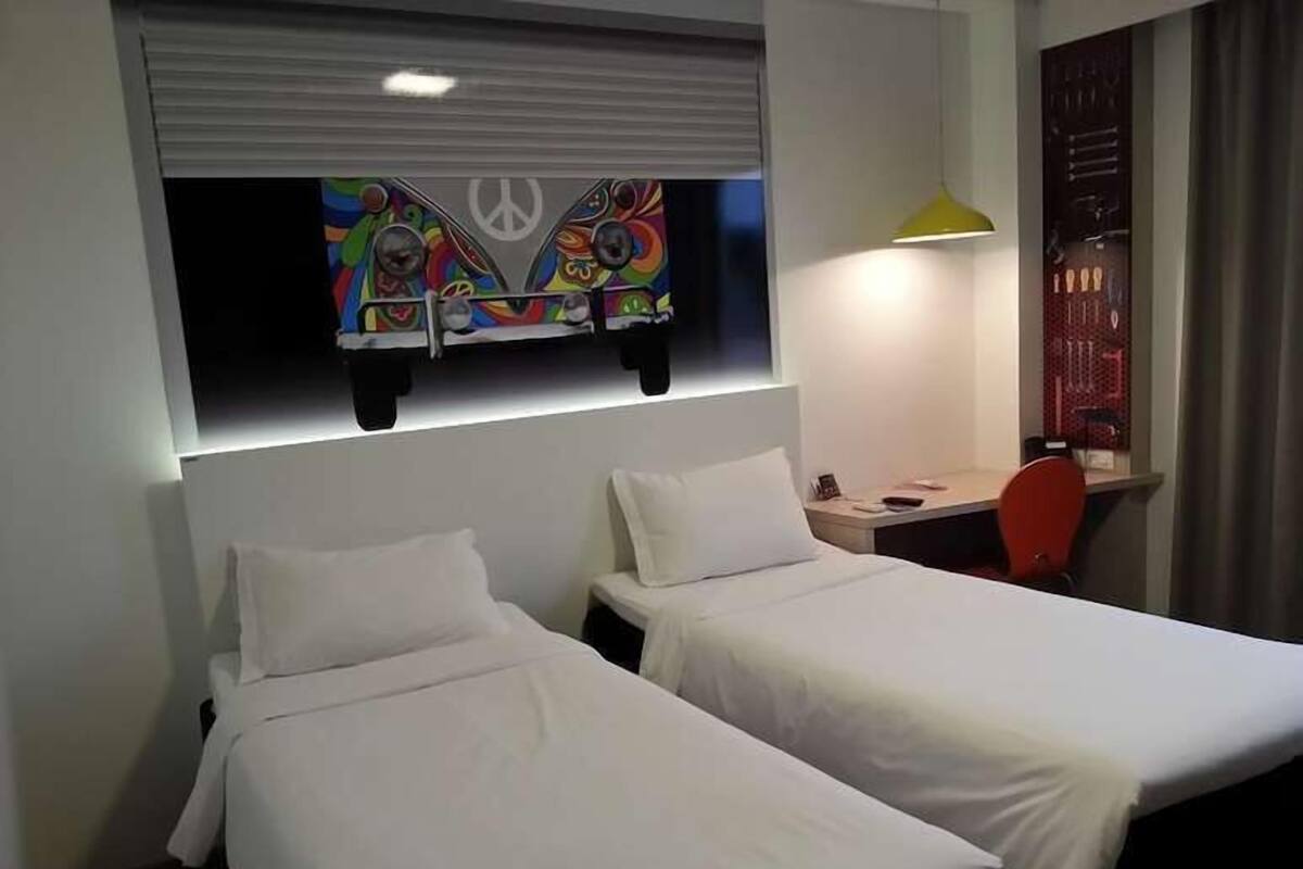 Habitación de hotel con dos camas individuales, un escritorio con una silla, una obra de arte colorida sobre la cama y una lámpara en el escritorio.