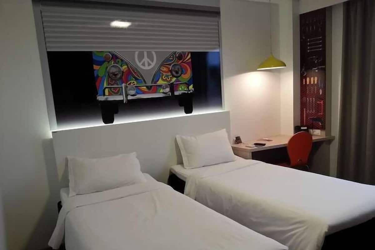 Una habitación de hotel con dos camas individuales, un escritorio, una silla, una obra de arte colorida en la pared y una lámpara.