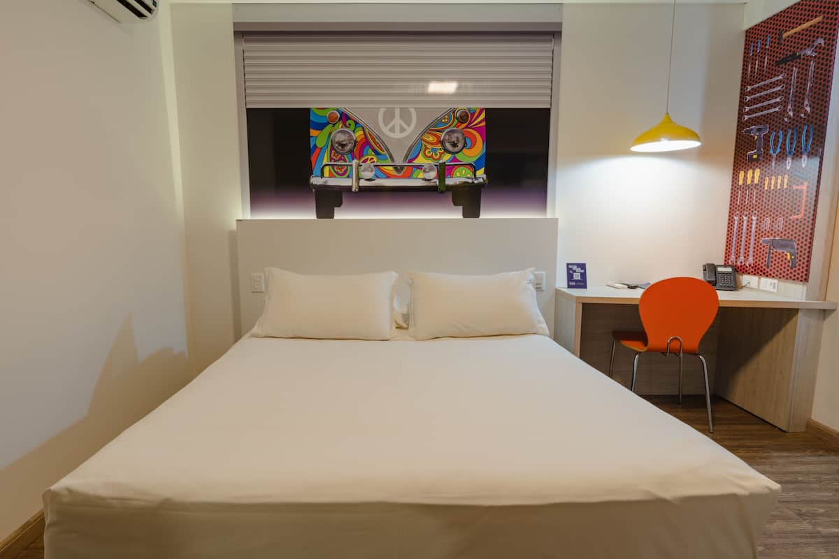 Un dormitorio con cama, un escritorio, una silla naranja y una obra de arte colorida sobre la cama.