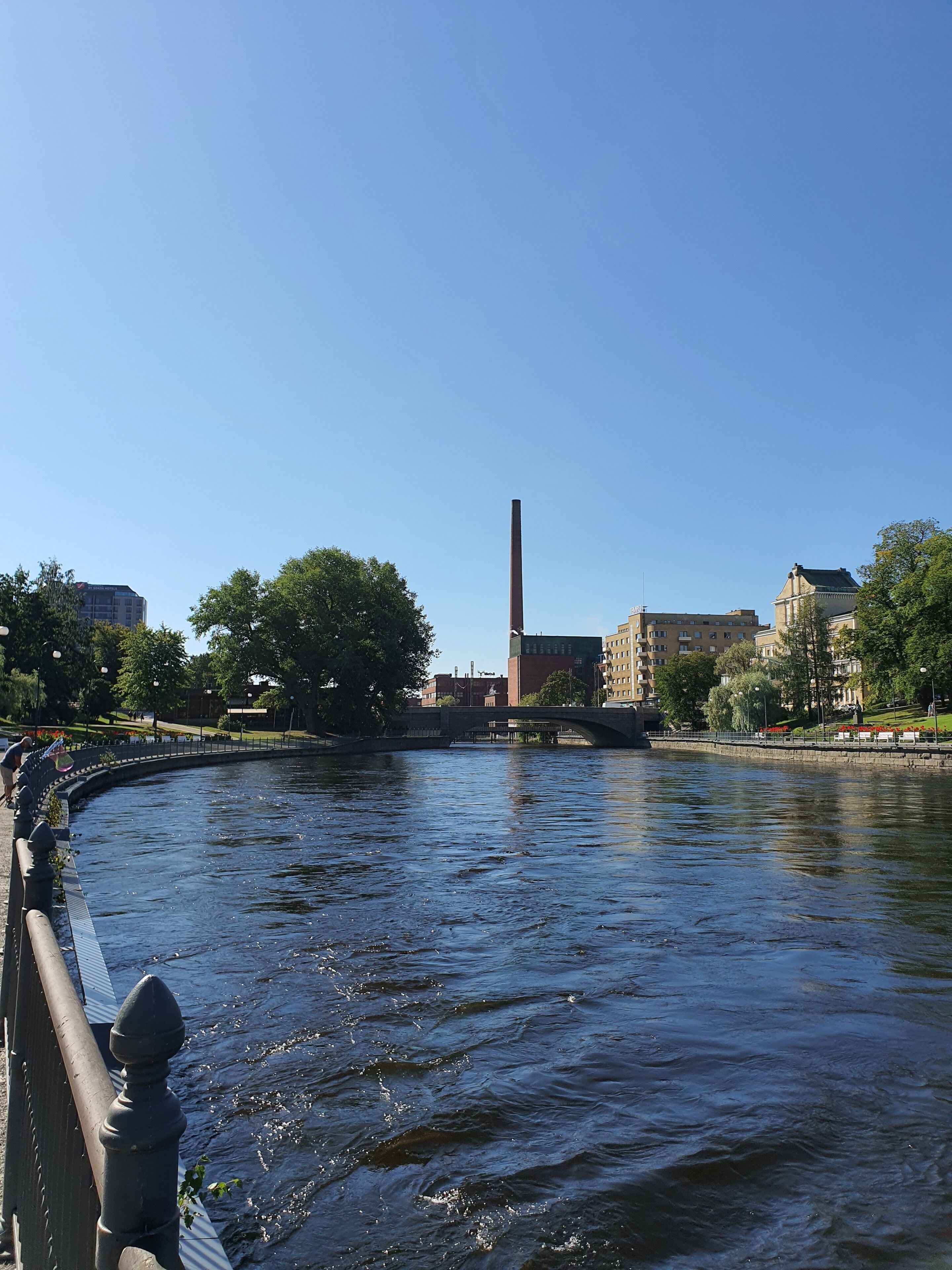 Une rivière avec un pont, des bâtiments et une haute cheminée en arrière-plan.