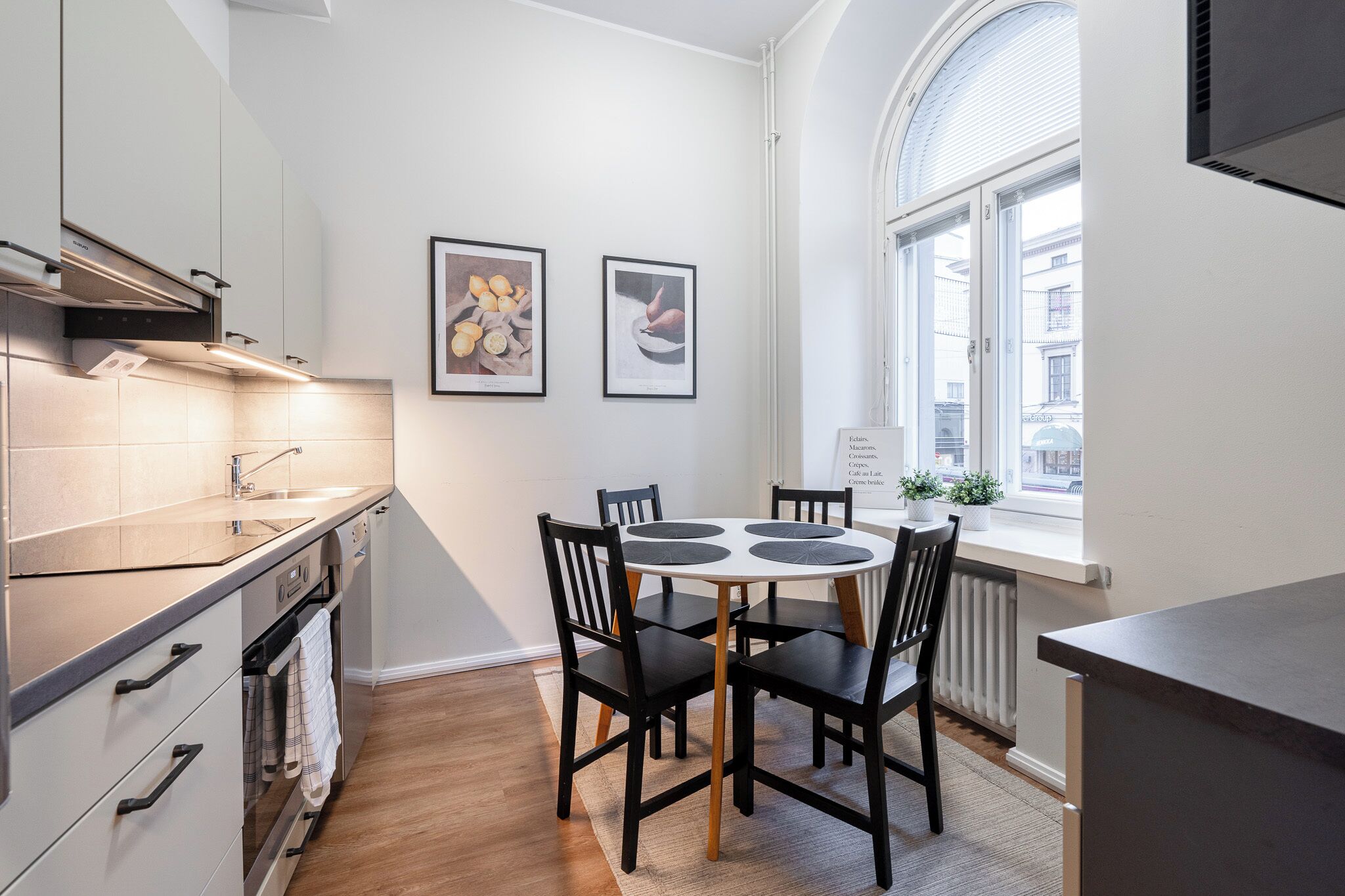 A modern kitchen with a dining area, featuring a round table with chairs, a built-in oven, and a window with a view of the street.