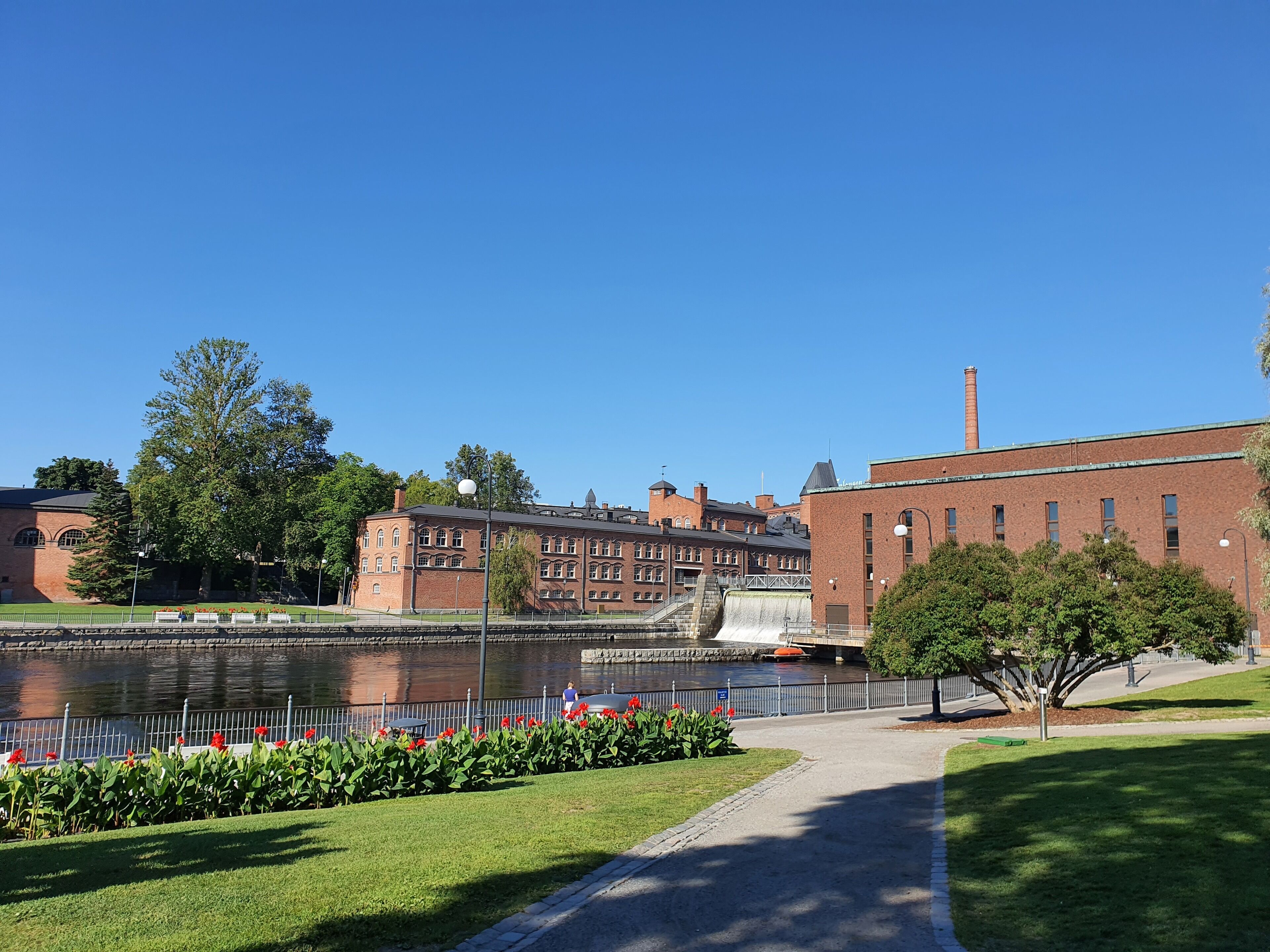A riverside walkway with a brick building, a bridge, and a water tower in the background.