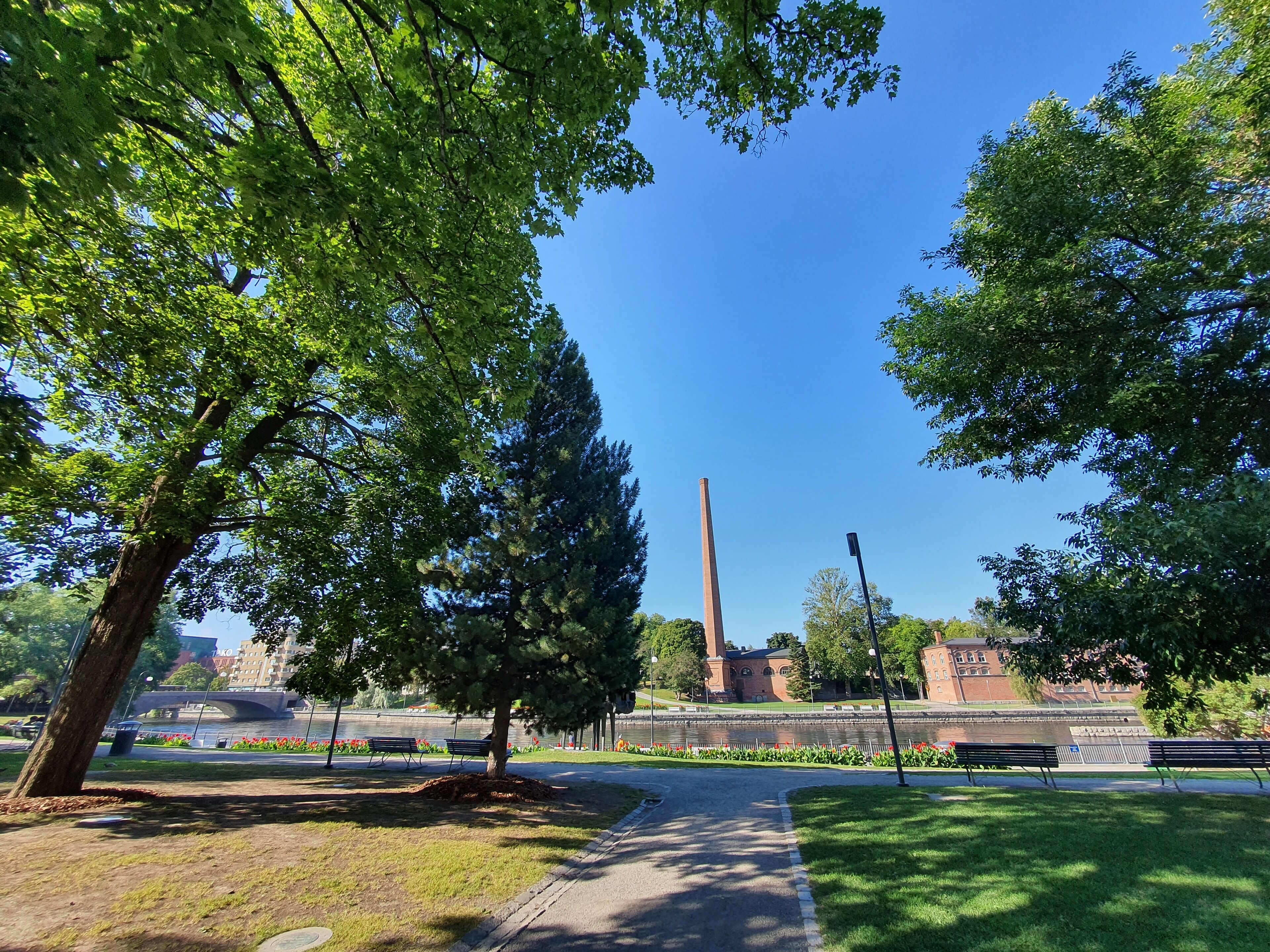 A park with a pathway, benches, trees, and a distant building with a chimney.