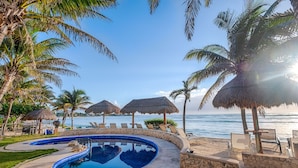 A pool area with a clear view of the ocean, surrounded by palm trees and thatched umbrellas. - Beautiful condo with ocean views, AC and pool! (Akumal)
