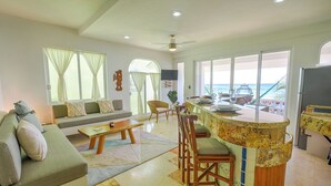 A spacious living area with a sofa, a coffee table, and a dining area with a bar. Large windows provide a view of the ocean. - Beautiful condo with ocean views, AC and pool! (Akumal)