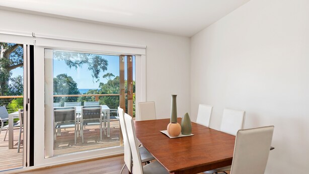 A dining area with a wooden table, white chairs, and a view through a glass door to an outdoor deck. - Strathlea (Lorne)