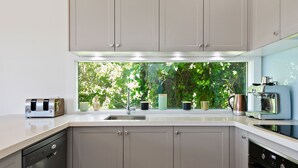 A modern kitchen with a built-in dishwasher, a coffee machine, and a window with a view of greenery. - Strathlea (Lorne)