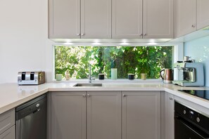 A modern kitchen with a built-in dishwasher, a coffee machine, and a window with a view of greenery. - Strathlea (Lorne)