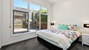 A bedroom with a bed, nightstands, a lamp, and a view through large windows. - Strathlea (Lorne)