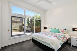 A bedroom with a bed, nightstands, a lamp, and a view through large windows. - Strathlea (Lorne)