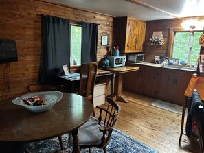 A wooden cabin interior with a dining table, chairs, a kitchen area with a microwave, and a window with curtains. - Gabriel`s Cabin in the Woods (Le Roy)