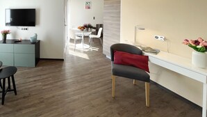 Un salon moderne doté d’un téléviseur à écran plat, d’un coin repas avec une table et des chaises blanches, et d’un espace de travail avec un bureau et une chaise.