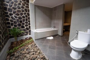 A bathroom with a stone wall, a bathtub, a toilet, and a small plant. - New Sunari Lovina Beach Resort (Buleleng)