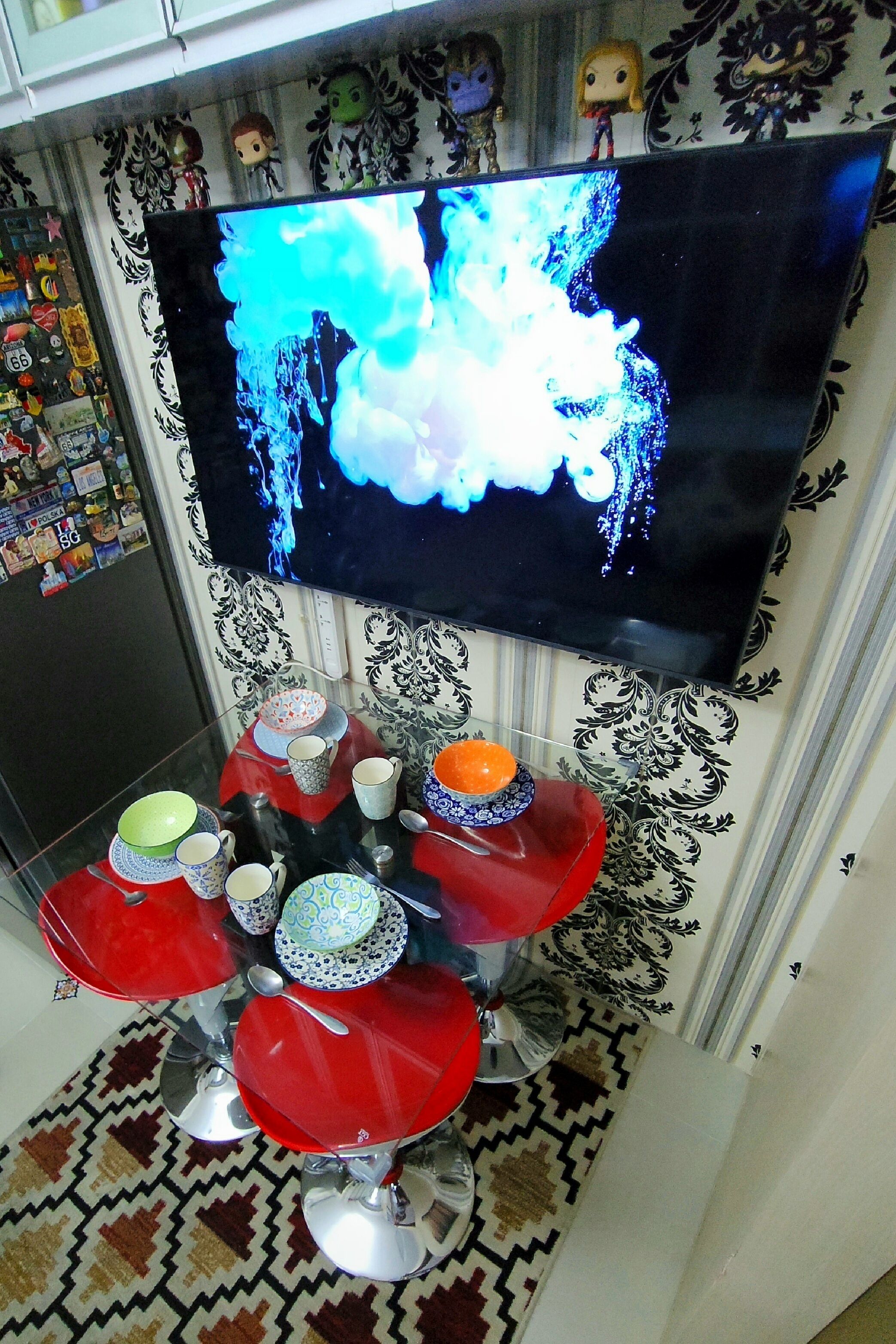 A room with a television mounted on the wall, a table with red chairs, and a patterned rug.