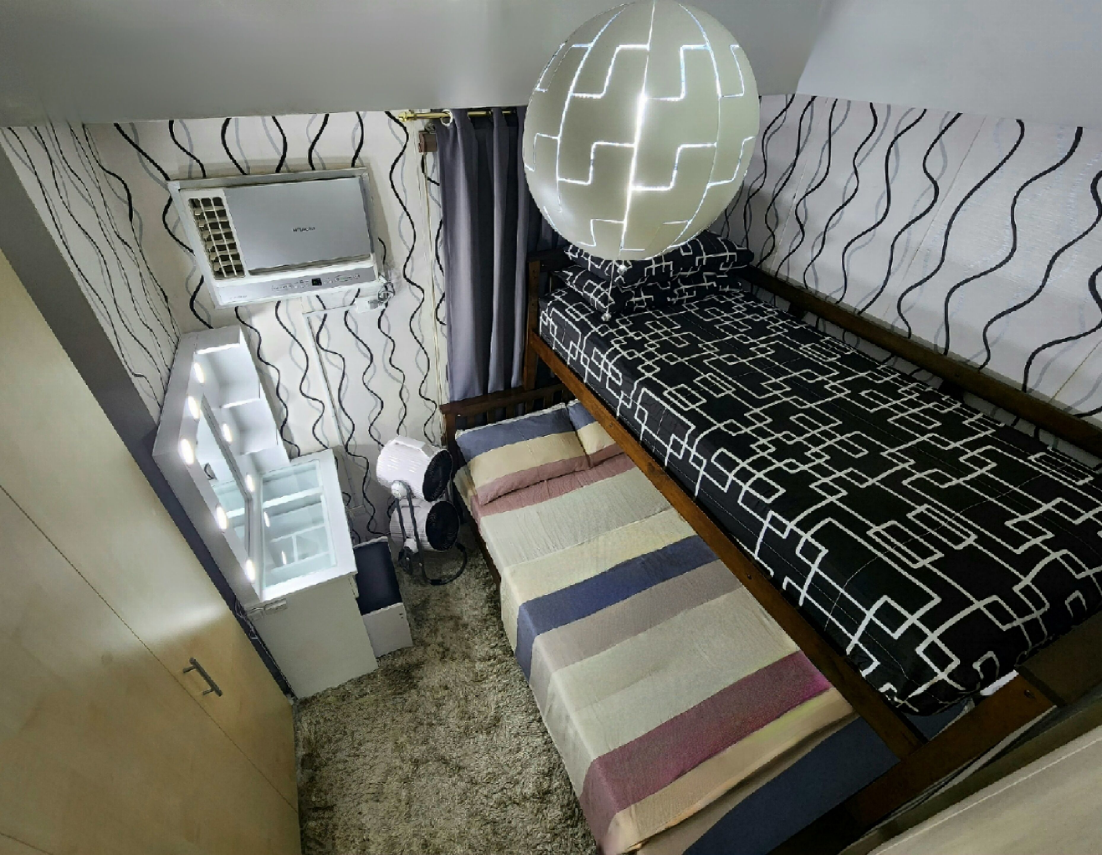 Una habitación pequeña con cama, tocador con luz, equipo de aire acondicionado y pared con diseño.