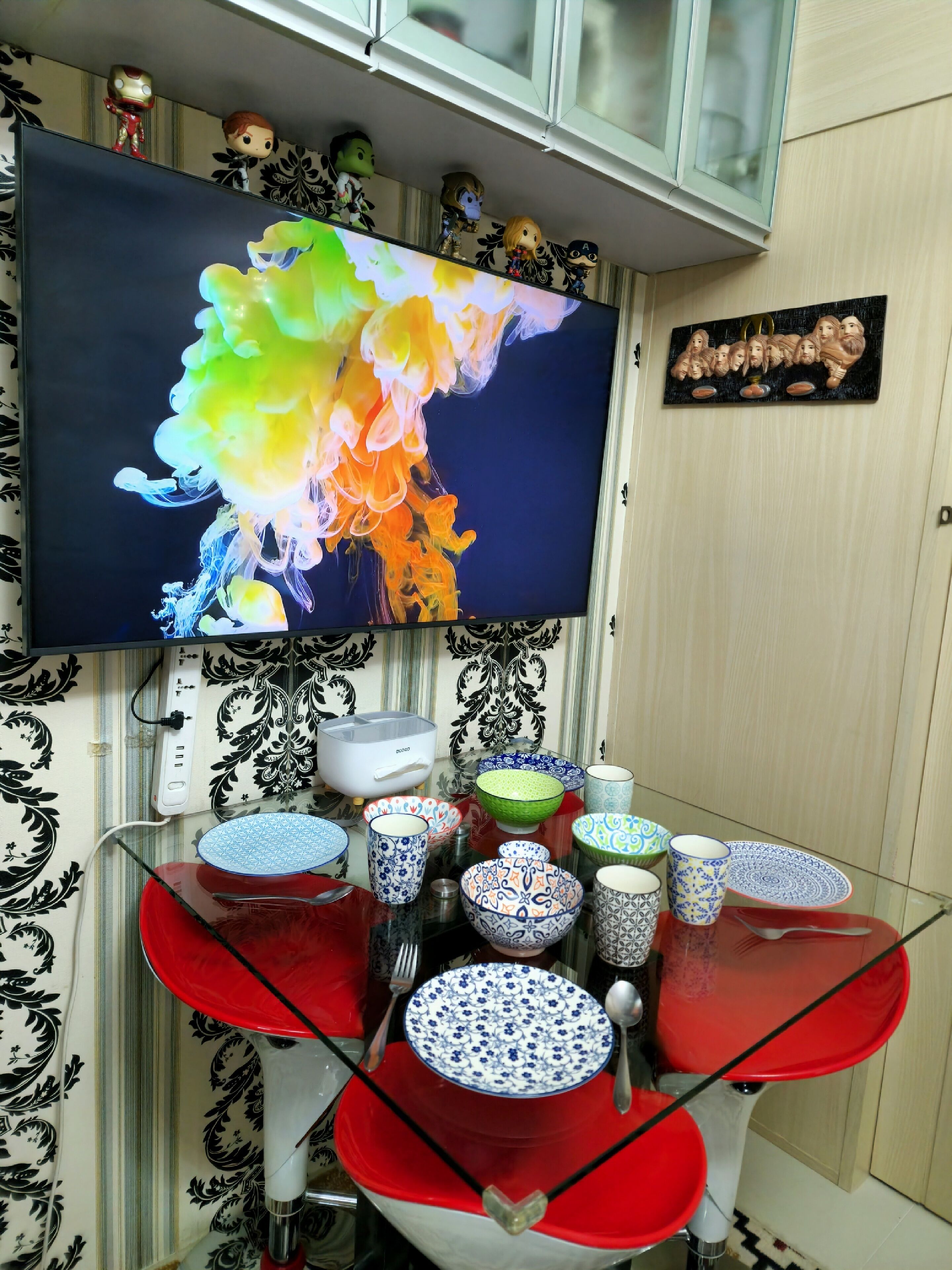 A kitchen with a TV displaying colorful abstract art, a variety of patterned bowls and cups on a glass table, and a decorative wall with patterned wallpaper and a small figurine collection.