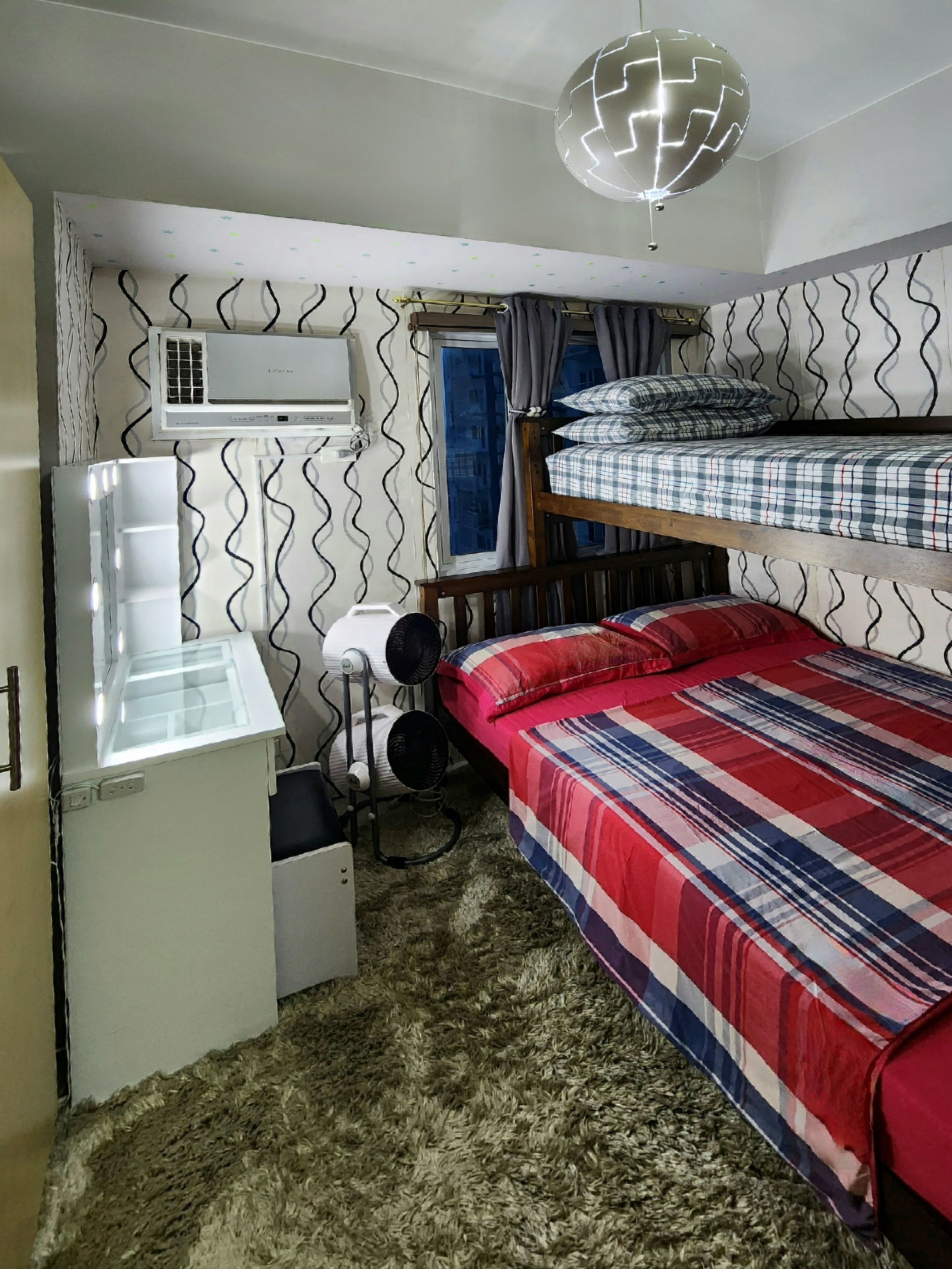 A compact room with bunk beds, a desk with a chair, a white refrigerator, a heater, and an air conditioning unit.
