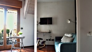 Uma sala de estar com televisão na parede, um sofá, uma mesa com uma planta e uma cadeira de balanço.