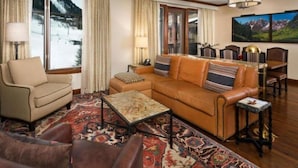 A cozy living room with a sofa, armchair, and coffee table, large windows with curtains, and a dining area with chairs and a chandelier. - Aspen Ritz-carlton 2 Bedroom Ski in, Ski out Residence With Access to Slopeside Heated Pools and Hot Tubs (Aspen)