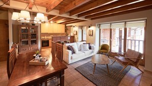 Un espacio habitable amplio con una mesa de comedor de madera, un sofá blanco y un sillón moderno. Se aprecia una cocina con armarios de madera y una zona de comedor con una araña en el fondo.