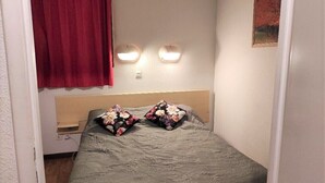 A small, cozy bedroom with a single bed, floral pillows, a red curtain, and two wall-mounted lights.