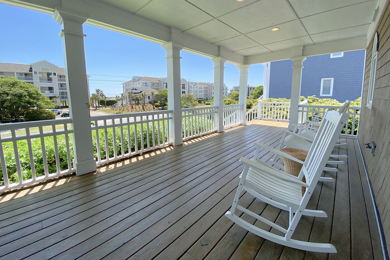 A covered porch with white railings and wooden flooring, overlooking a residential area with houses and greenery.