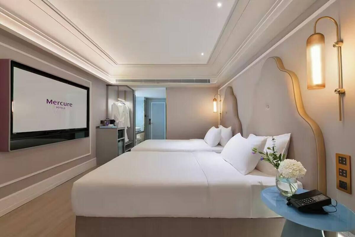 A hotel room with a bed, bedside table, lamp, and a screen displaying 'Mercure Hotels'.
