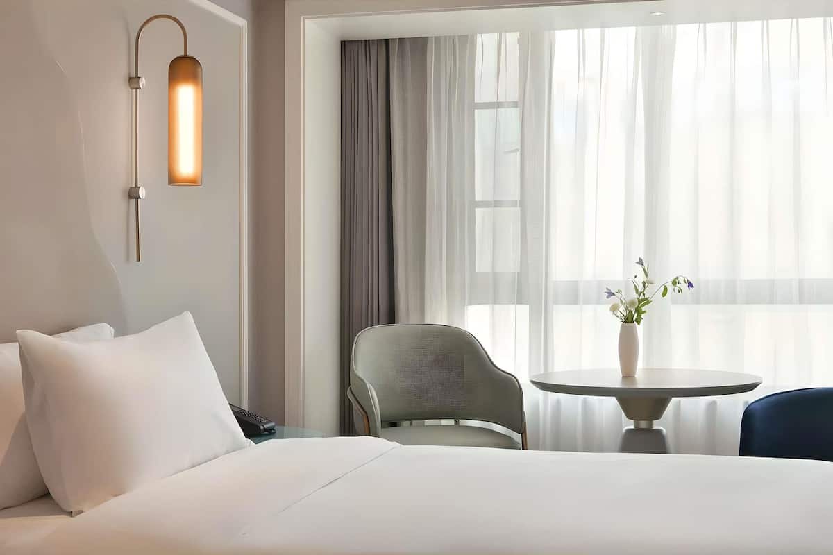 A hotel room with a bed, a chair, a small table with a vase, and a wall-mounted light fixture.