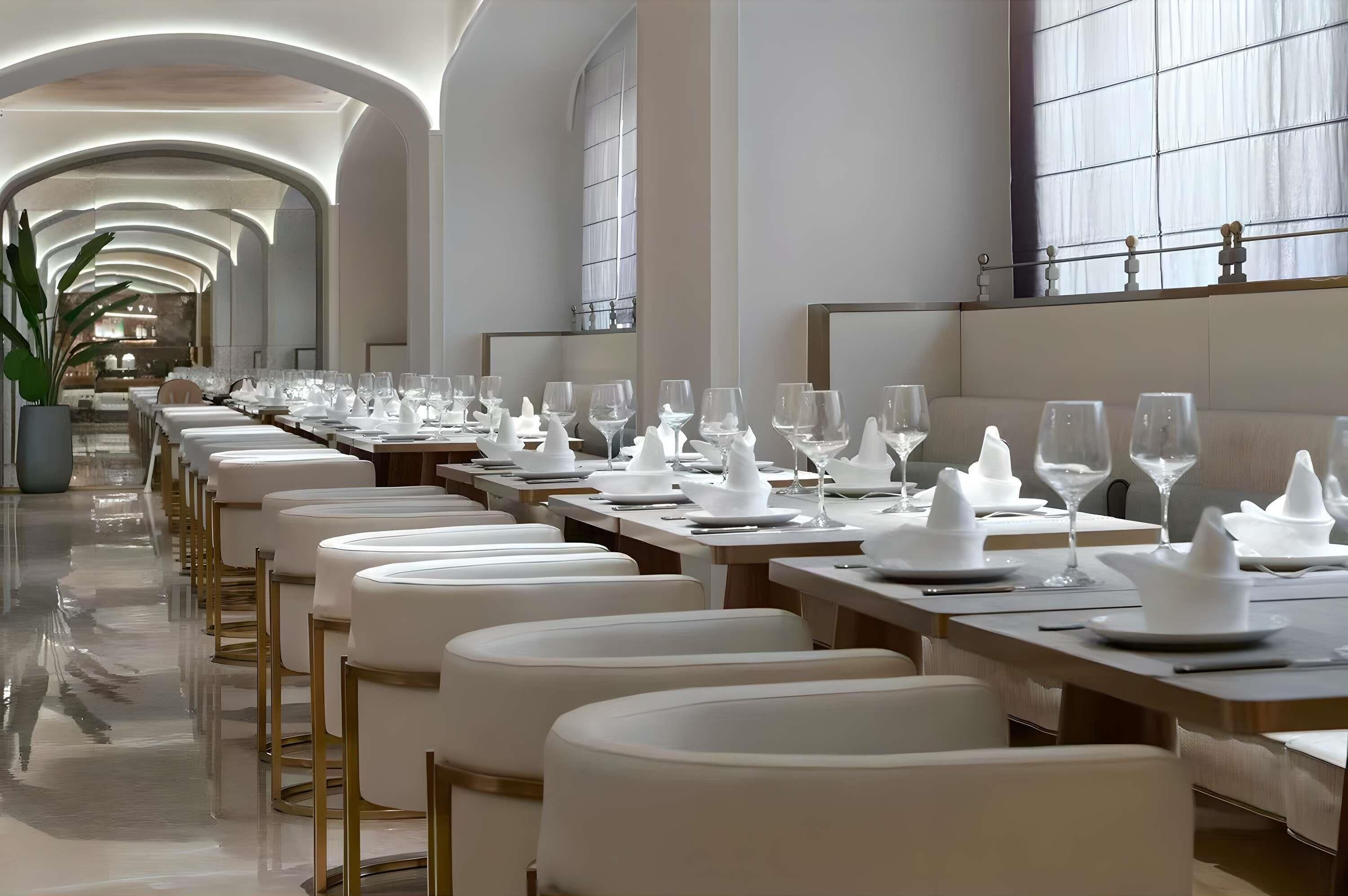 A long dining table with white tablecloths, chairs, and glassware, set in a well-lit, modern interior with arched ceilings and large windows.