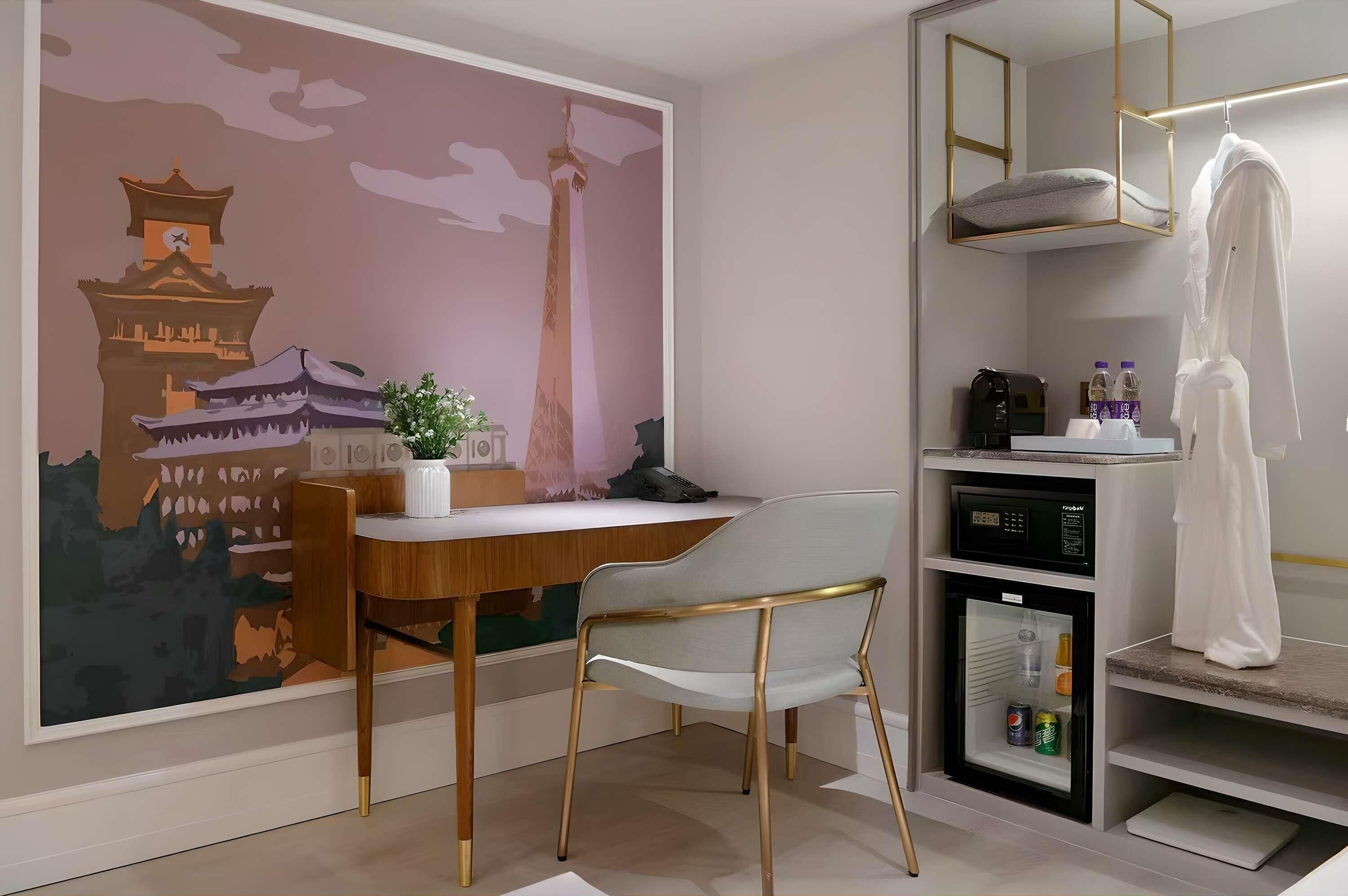 Ein Hotelzimmer mit Schreibtisch aus Holz, einem weißen Stuhl, einer Minikühlschrank, einer kleinen Vitrine, einem Spiegel und einer Wand mit einem Wandgemälde eines traditionellen Gebäudes.