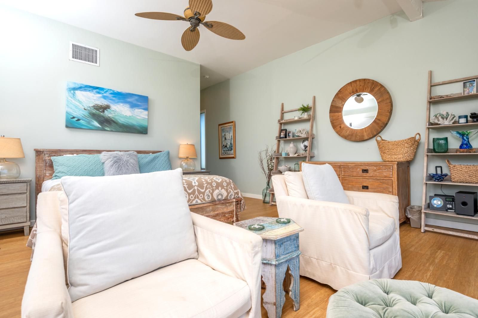 A spacious living room with a large bed, two armchairs, a coffee table, and a wooden dresser. There is a ceiling fan, a painting, and a mirror on the wall.
