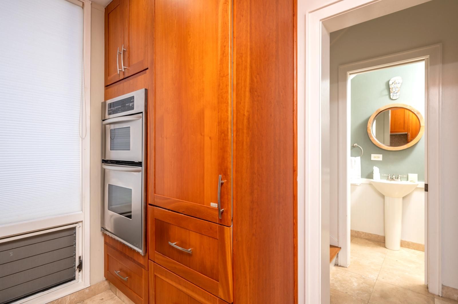 A kitchen with wooden cabinets, a built-in oven, and a view into the adjacent bathroom with a sink and mirror.