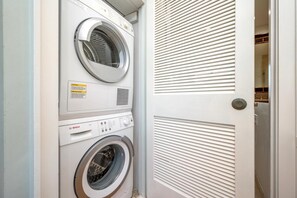 Une machine à laver et un sèche-linge superposés dans une buanderie, avec une porte donnant sur un couloir.