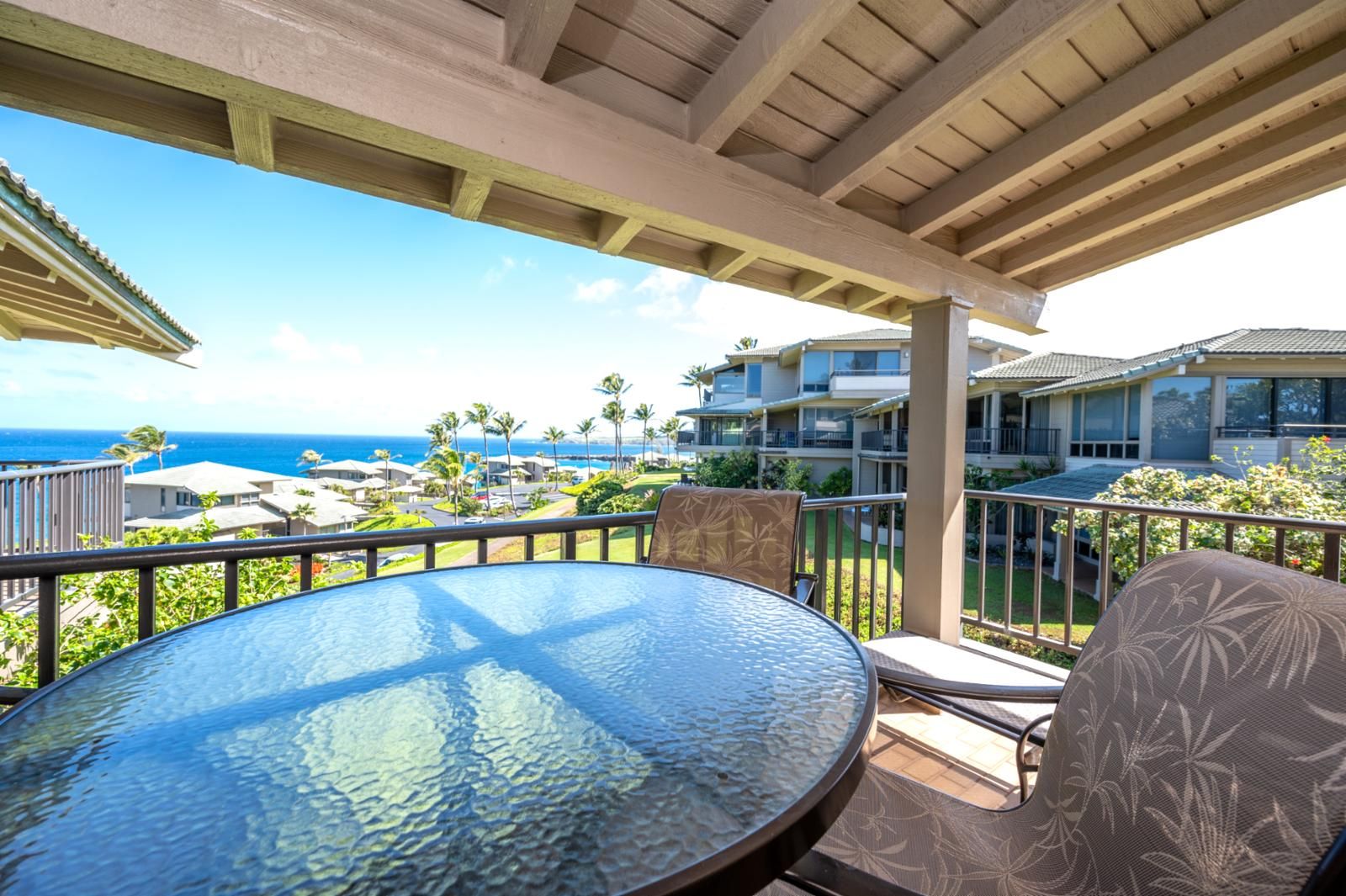 A covered outdoor area with a round glass table and two chairs, overlooking a residential area with a view of the ocean.