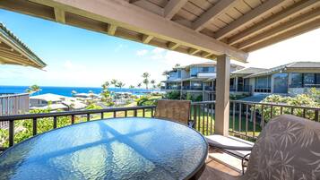 A covered outdoor area with a round glass table and two chairs, overlooking a residential area with a view of the ocean.