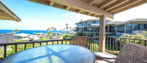 A covered outdoor area with a round glass table and two chairs, overlooking a residential area with a view of the ocean.