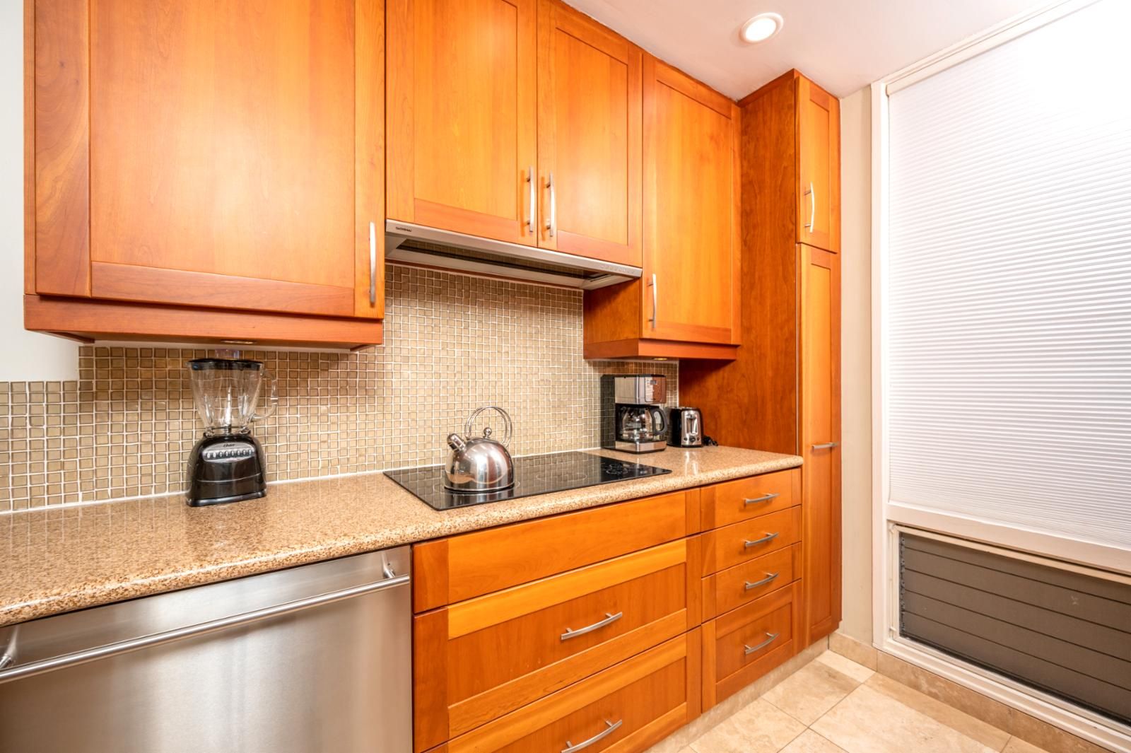 A kitchen with wooden cabinets, a stainless steel dishwasher, a blender, a kettle, and a coffee maker on a tiled backsplash.