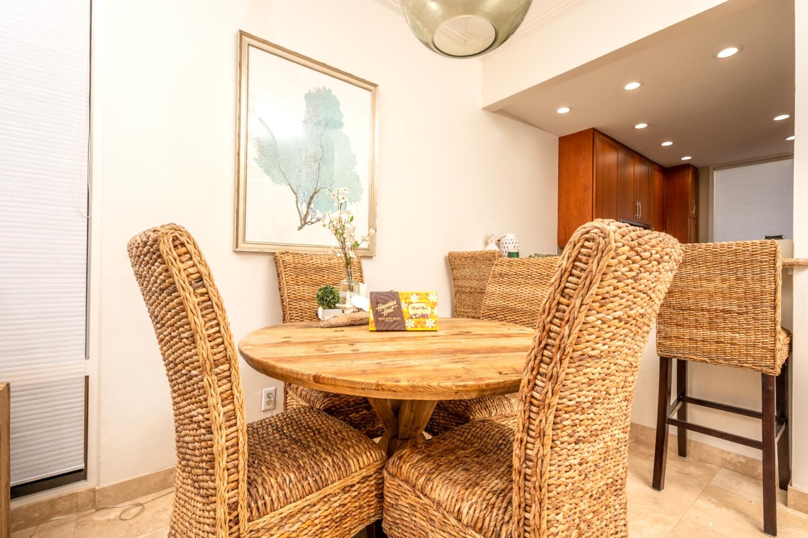 A dining area with a round wooden table surrounded by woven chairs, a framed artwork on the wall, and a kitchen in the background.