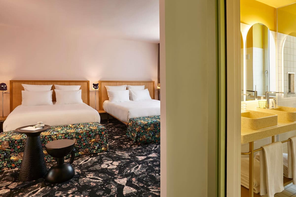 A hotel room with two beds, a small table, a patterned ottoman, and a bathroom with a sink and mirror.