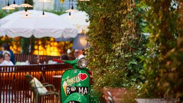 En grøn scooter parkeret i et udendørs område med borde og stole, omgivet af grønt og hængende lys.