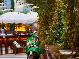 Uno scooter verde parcheggiato in un'area esterna con tavoli e sedie, circondato dal verde e da luci appese.
